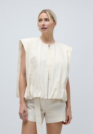 Blondynka w bezrękawnym, kremowym, pikowanym topie na zamek błyskawiczny i dopasowanych szortach, stojąca na tle jasnoszarego tła, patrząca w lewo.