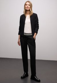 Schwarze Bomberjacke über einem cremefarbenen Oberteil mit gestreiftem Bund. Schwarze Hose mit Taschen und schwarze Stiefeletten. Einfaches und modernes Design.