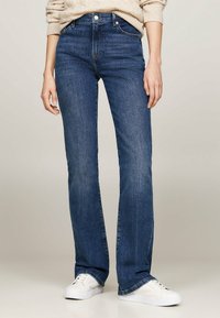 Pantalones vaqueros oscuros de tiro acampanado, confeccionados en denim, con cintura alta, diseño de cinco bolsillos y sutiles detalles de costura. Combinados con zapatillas blancas.