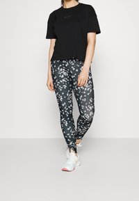 Černé tričko s krátkým rukávem v kombinaci s přiléhavými legínami s tmavým pozadím a bílým abstraktním květinovým vzorem, doplněné o bílé tenisky.