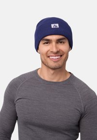 Navy knit beanie with a folded cuff and a small white logo patch; worn by a person in a gray long-sleeve shirt. Smooth texture.