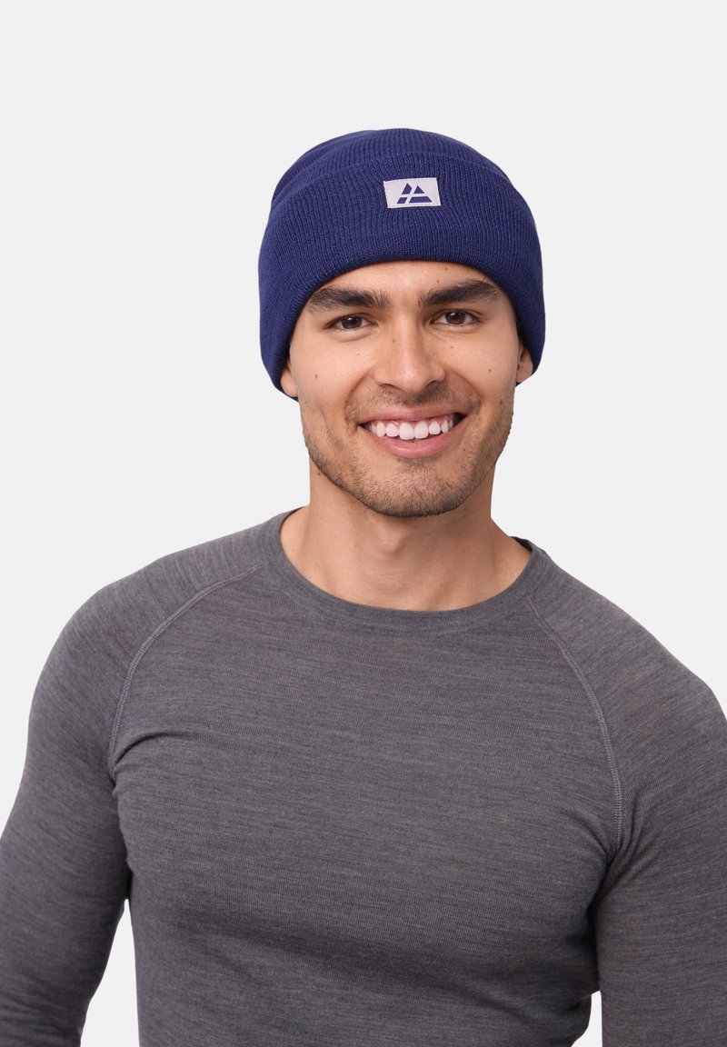 Navy knit beanie with a folded cuff and a small white logo patch; worn by a person in a gray long-sleeve shirt. Smooth texture.