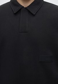 Chemise noire à col avec une courte patte de boutonnage et une petite pièce rectangulaire avec texte en relief en bas à droite.