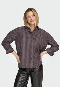 Camisa de cuadros abotonada en rojo oscuro y azul marino, de corte holgado, mangas largas, bolsillo en el pecho, combinada con pantalones de cuero negro brillante.