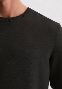 Gros plan sur un pull en maille noir à col rond porté par une personne, mettant en évidence un logo embossé subtil au niveau de la poitrine.