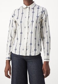 Person trägt ein weißes Langarmhemd mit blauen vertikalen Streifen und kreisförmigen Mustern, kombiniert mit dunkelblauen Jeans.