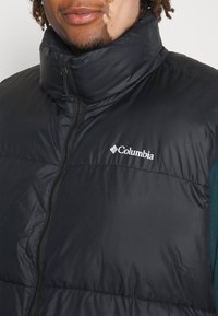 Chaleco aislante negro con cuello alto, cierre con cremallera y logo de Columbia. Textura lisa y brillante con un patrón sutil y detalles de costura.