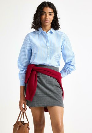 Lys blå skjorte med hvide striber, parret med en grå ternet nederdel og en kastanjebrun sweater bundet om taljen. Brun taske i hånden.