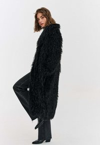 Žena v dlouhém černém plyšovém kabátu, černých džínách a černých kozačkách na podpatku, stojící s jednou nohou zdviženou na jednoduchém bílém pozadí.