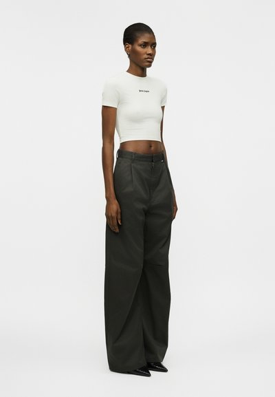 Γυναίκα όρθια, φοράει λευκό cropped t-shirt με κείμενο και μαύρο παντελόνι φαρδύ με μαύρα μυτερά παπούτσια, κοιτάζει δεξιά.