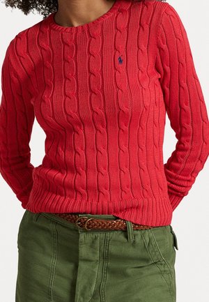 Personne portant un pull rouge à torsades avec un petit logo brodé et un pantalon vert olive avec une ceinture tressée marron.