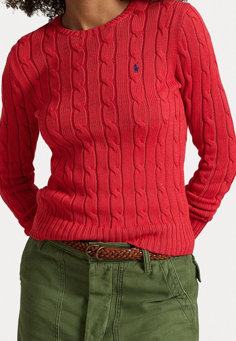 Personne portant un pull rouge à torsades avec un petit logo brodé et un pantalon vert olive avec une ceinture tressée marron.