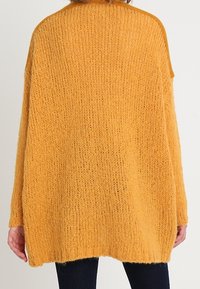 Oversized yellow knit sweater made of textured yarn, featuring long sleeves and a rounded hem, displayed from the back.
