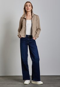 Veste zippée beige avec deux poches avant, débardeur blanc et jeans larges bleu foncé. Portée avec des baskets blanches sur un fond gris.