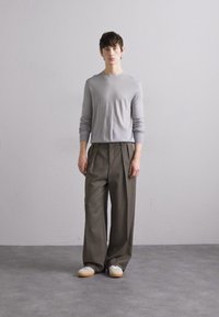 Chemise ajustée à manches longues gris clair associée à un pantalon large marron foncé et des baskets blanches avec des semelles beiges, se tenant sur un sol gris.