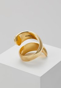Gold-tone metal ring with a smooth, curved design featuring two overlapping loops. Engraved branding on the interior.