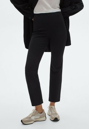 Osoba ubrana w czarne spodnie do połowy łydki, beżowo-białe sneakersy, biały top i czarną kurtkę, z rękami w kieszeniach.