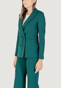 Modelo vistiendo un blazer verde de doble botonadura con pantalones a juego, mostrando un atuendo profesional contra un fondo gris claro.