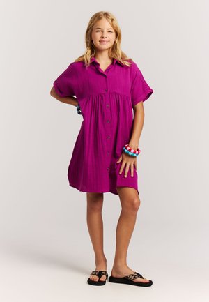 Fuchsia blousejurk met korte mouwen, een ingezette taille en een losse pasvorm. Draagt zwarte slippers en kleurrijke armbanden om één pols.