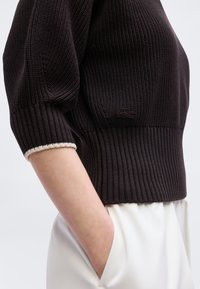 Πλάγια όψη ατόμου που φοράει πλεκτό σκούρο καφέ πουλόβερ με μανίκια τριών τετάρτων και λευκό παντελόνι, το χέρι στην τσέπη.