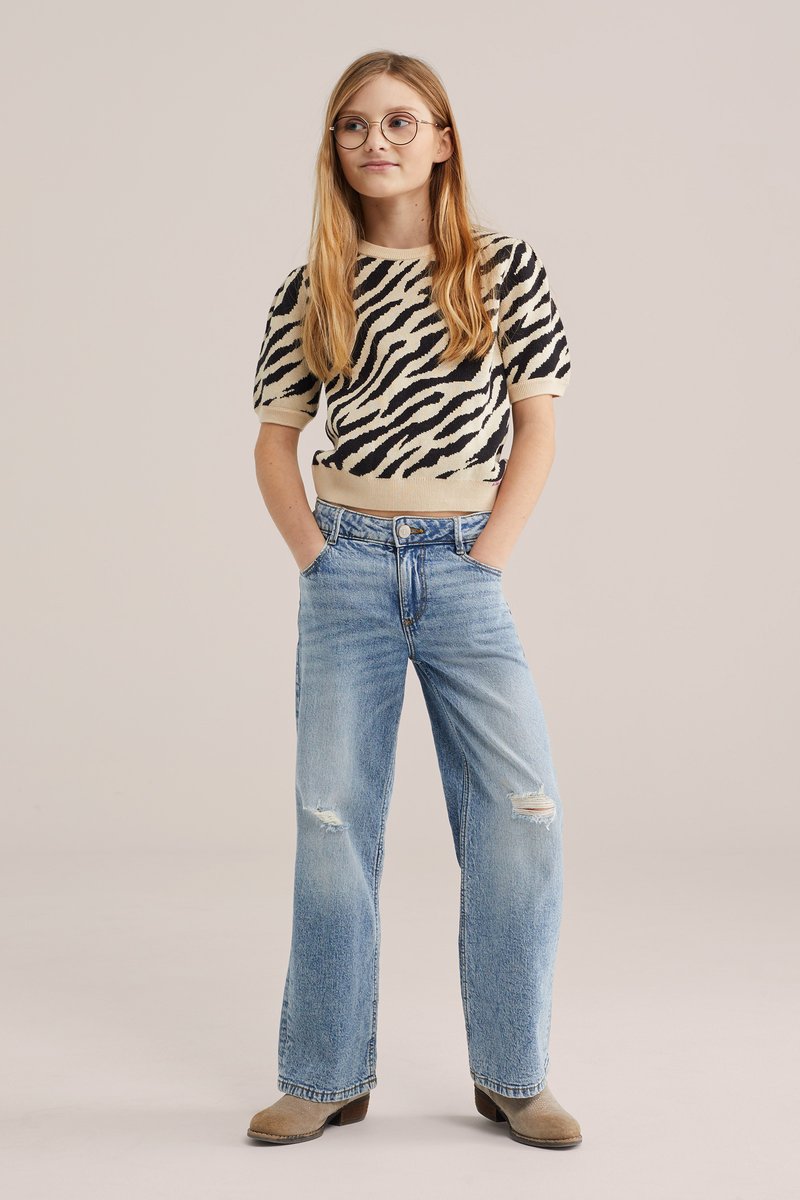 Jong persoon met lang blond haar, rond brilletje, zebraprint kortemouwen shirt, lichtblauwe gescheurde spijkerbroek en beige laarzen, staand met handen in de zakken.