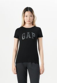Woman standing, wearing a black GAP t-shirt and dark pants, facing forward against a plain white background.