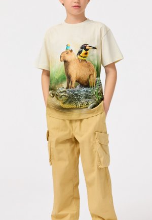 Maglietta beige chiaro con una grafica di un capibara, uccelli e un coccodrillo, su uno sfondo verde. Abbinata a pantaloni cargo gialli.
