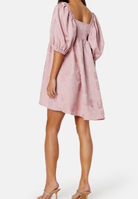 Vaaleanpunainen, tekstuurilla varustettu mekko puffihousuilla ja neliömäisellä selällä. Kankaan pinnassa on hienovarainen kukkakuosi. Yhdistetty nudet korkokengät.