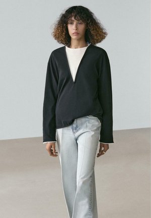 Giovane donna con capelli ricci che indossa una maglia nera a maniche lunghe sopra una camicia bianca e jeans larghi azzurri chiari, in piedi all'interno.