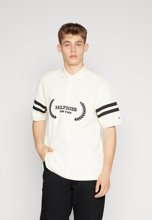 Polo bianco con strisce nere sulle maniche e logo ricamato "HILFIGER NEW YORK" circondato da motivi di alloro. Indossato con pantaloni neri.