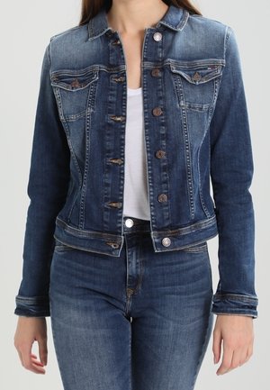 Kvinde iført en tætsiddende blå denimjakke over en hvid skjorte med matchende blå denimjeans, stående med arme afslappet langs siderne.