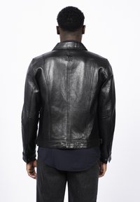 Chaqueta de cuero negra, diseño ajustado, textura suave, cuello ancho y detalles de costura visibles. Presenta un parche con el logo en la parte posterior.