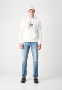 Hvit hettegenser med logo og marineblå detaljer, blå jeans og svarte sko. Modellen har på seg en lyse farget caps. Enkelt, uformelt antrekk.