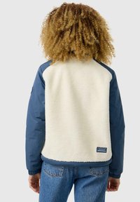 Fleece in najlonska jakna s krem telesom in mornarsko modrimi rokavi. Ima rebraste manšete in logo našitek na robu. Zgornji del je teksturiran, notranjost pa je mehka.
