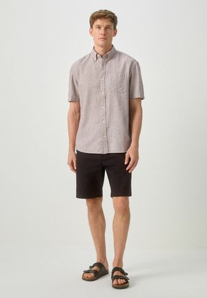 Man wearing short-sleeve button-up shirt, black shorts, and black buckle sandals standing against plain background.