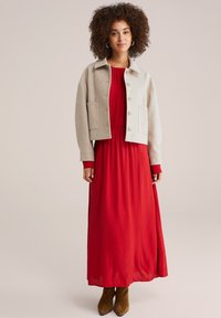 Grå uldblandet cropped jakke med fire knapper, parret med en flydende rød maxi kjole og brune ankelstøvler. Modellen står foran en neutral baggrund.