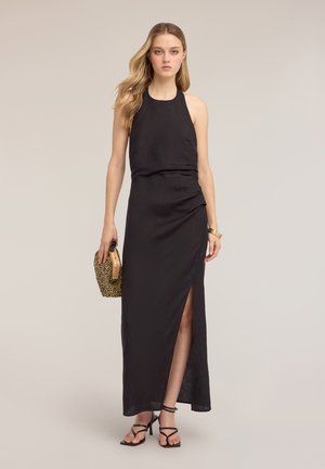 Mujer con un vestido maxi negro sin mangas con abertura lateral, sosteniendo un pequeño bolso tejido, con una pulsera dorada y tacones negros con tiras.
