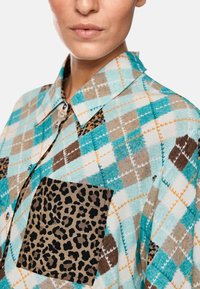 Checkered shirt with turquoise, brown, and white patterns. Features a leopard print pocket. Soft fabric with a classic collar and buttons.