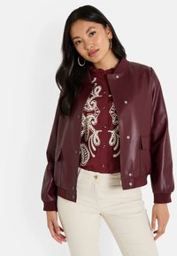 Chaqueta bomber de imitación piel burdeos con botones a presión, puños acanalados y dos bolsillos delanteros, superpuesta a una blusa de color burdeos con estampado.