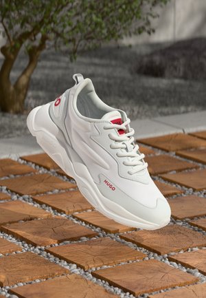 White sneaker featuring textured synthetic material, lace-up closure, red accents with branding, and a chunky sole, set against wooden tiles.