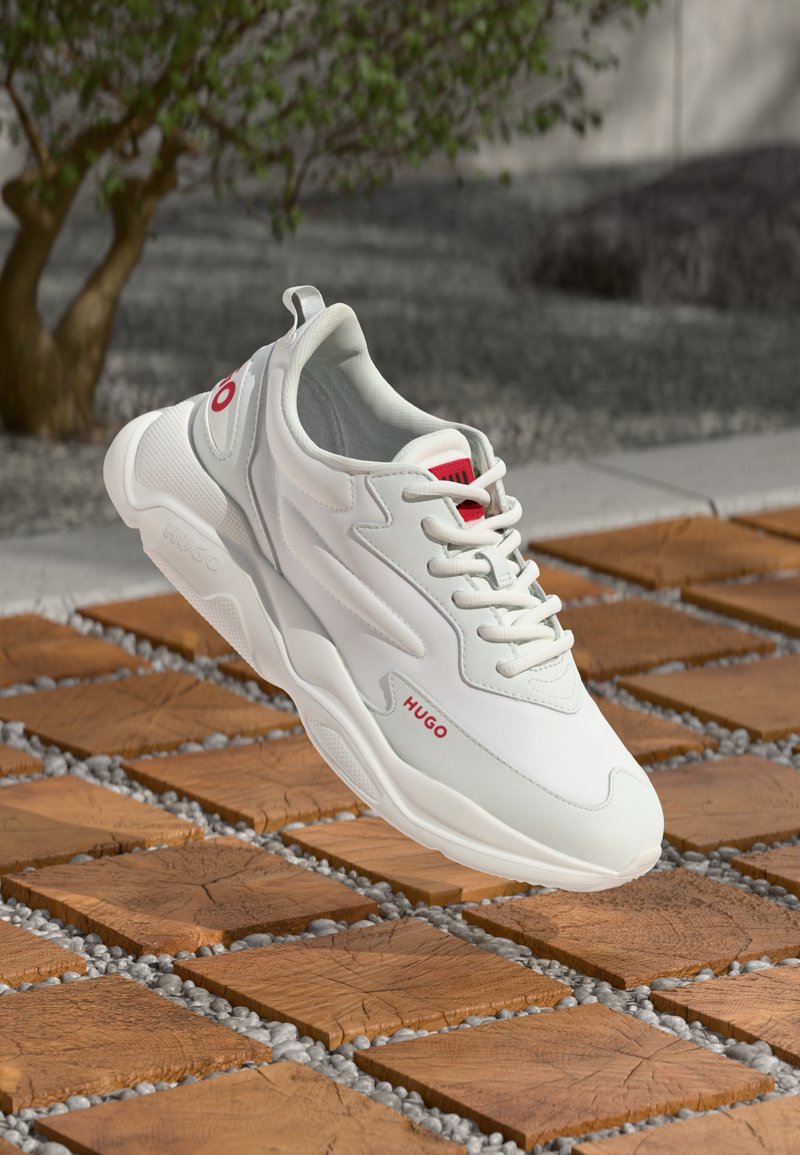 White sneaker featuring textured synthetic material, lace-up closure, red accents with branding, and a chunky sole, set against wooden tiles.