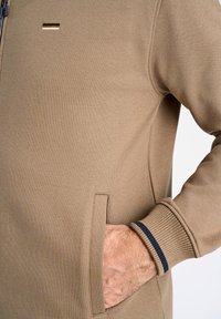 Felpa beige con zip, dotata di tasche laterali, polsini a coste con accenti blu navy, tessuto testurizzato e un piccolo logo orizzontale sul petto.