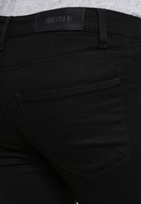 Jeans de denim negro con una vista cercana del bolsillo trasero y la cinturilla que presenta la marca "WHYRED" grabada en piel. Textura suave.