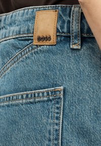 Κοντινό της πίσω τσέπης μπλε τζιν με καφέ δερμάτινο ταμπελάκι με την ένδειξη «BOSS» ραμμένο κοντά στη μέση.