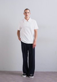 Homme se tenant contre un mur simple portant un polo blanc à manches courtes, un pantalon large noir et des baskets noires avec des semelles blanches.