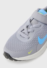 Baskets grises avec tige en mesh, logo en accent bleu et détails jaunes. Présente des lacets et une sangle Velcro. Semelle en caoutchouc texturée.