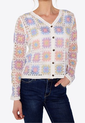 Kobieta w szydełkowanym kardiganie z pastelowymi wzorami z kwadratów granny w różnych kolorach i ciemnych dżinsach, z ręką w kieszeni.