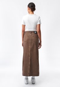 Falda maxi de denim marrón con bolsillos traseros, cintura alta y una costura visible en el centro, combinada con una blusa blanca de manga corta y zapatillas blancas.