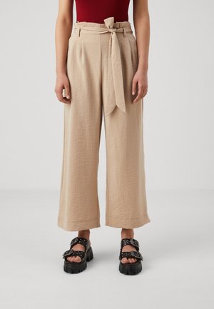 Frau trägt beige, hoch taillierte, weit geschnittene Hose mit gebundenem Gürtel, rotes ärmelloses Oberteil und schwarze klobige Sandalen mit Schnallen.