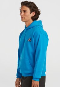 Sweat à capuche bleu en matière polaire. Doté d'une poche kangourou à l'avant et d'un patch logo triangulaire sur la poitrine.